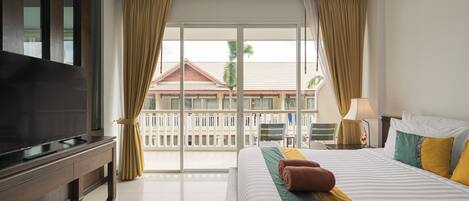 A hotel room with a large flat-screen TV, a bed with striped bedding, a green and yellow throw blanket, and a view of a building with a red roof through the curtains.