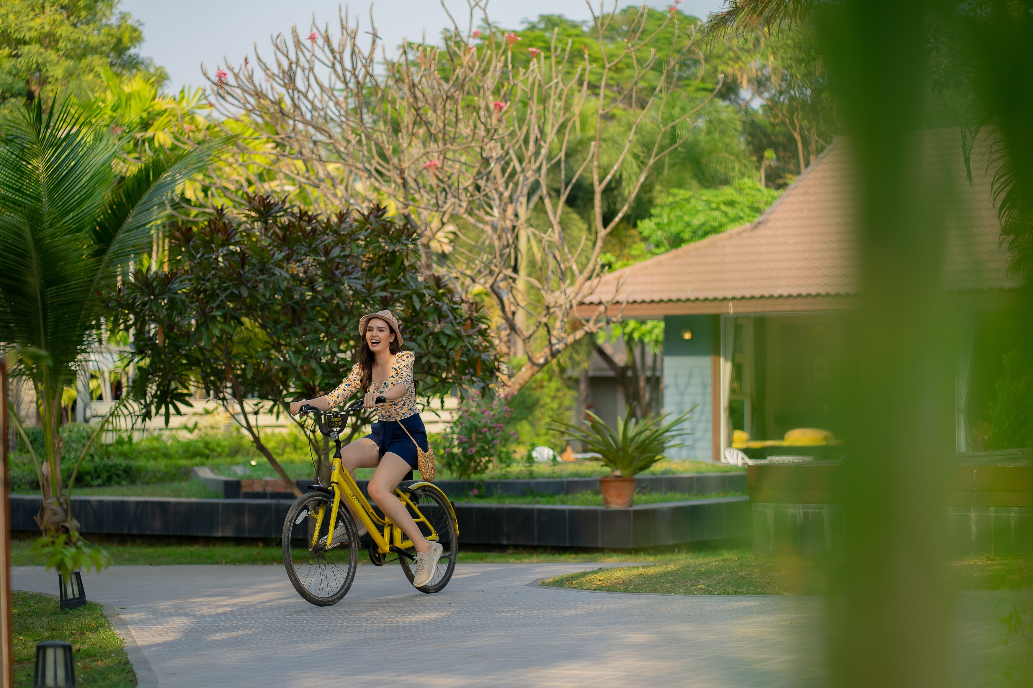 Una mujer pedaleando una bicicleta amarilla por un camino pavimentado en un exuberante jardín con diversas plantas y árboles.