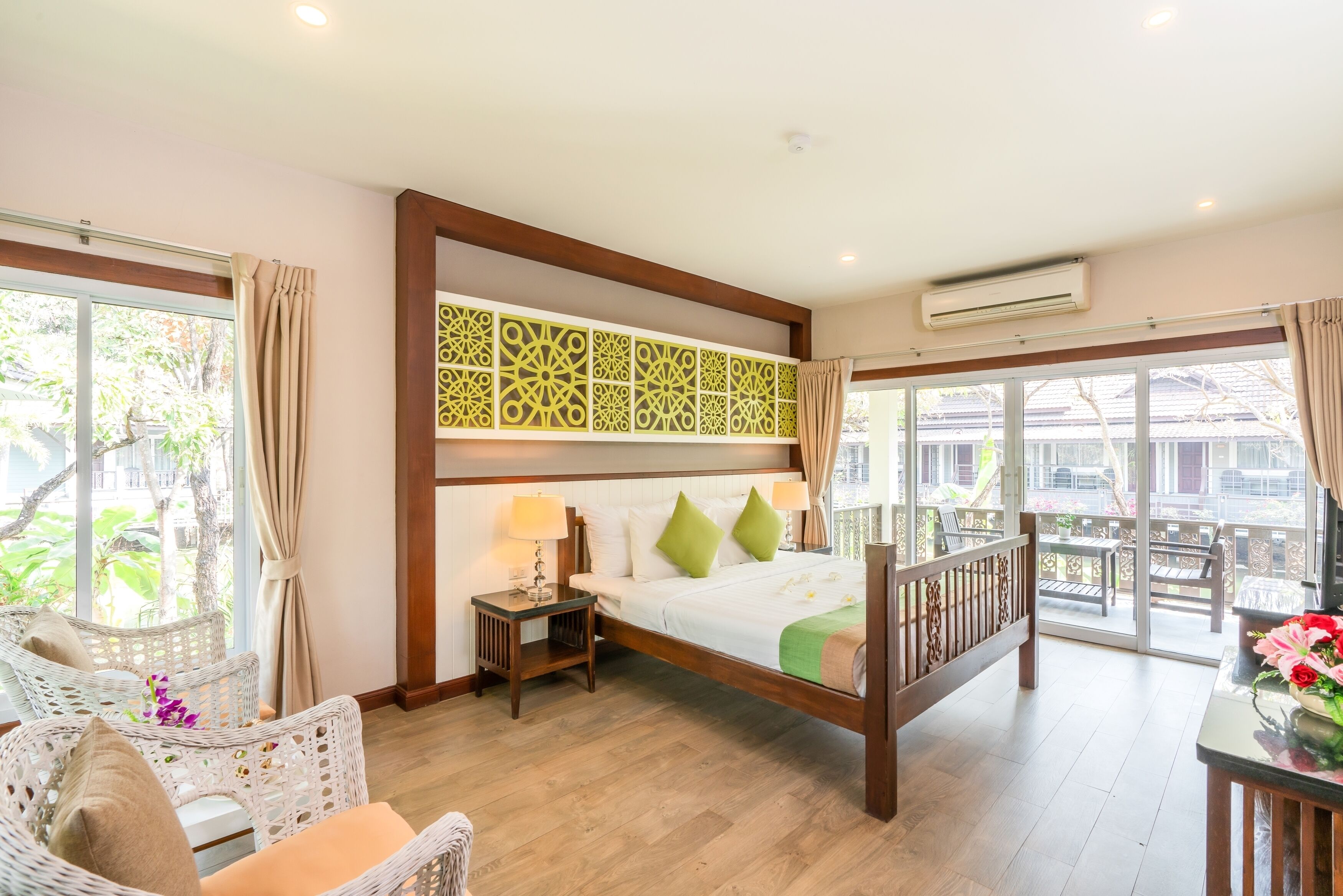 A hotel room with a bed, a chair, a table with flowers, and a balcony with outdoor furniture.