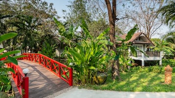 A wooden bridge with red railings, surrounded by lush greenery and a house with a tiled roof.