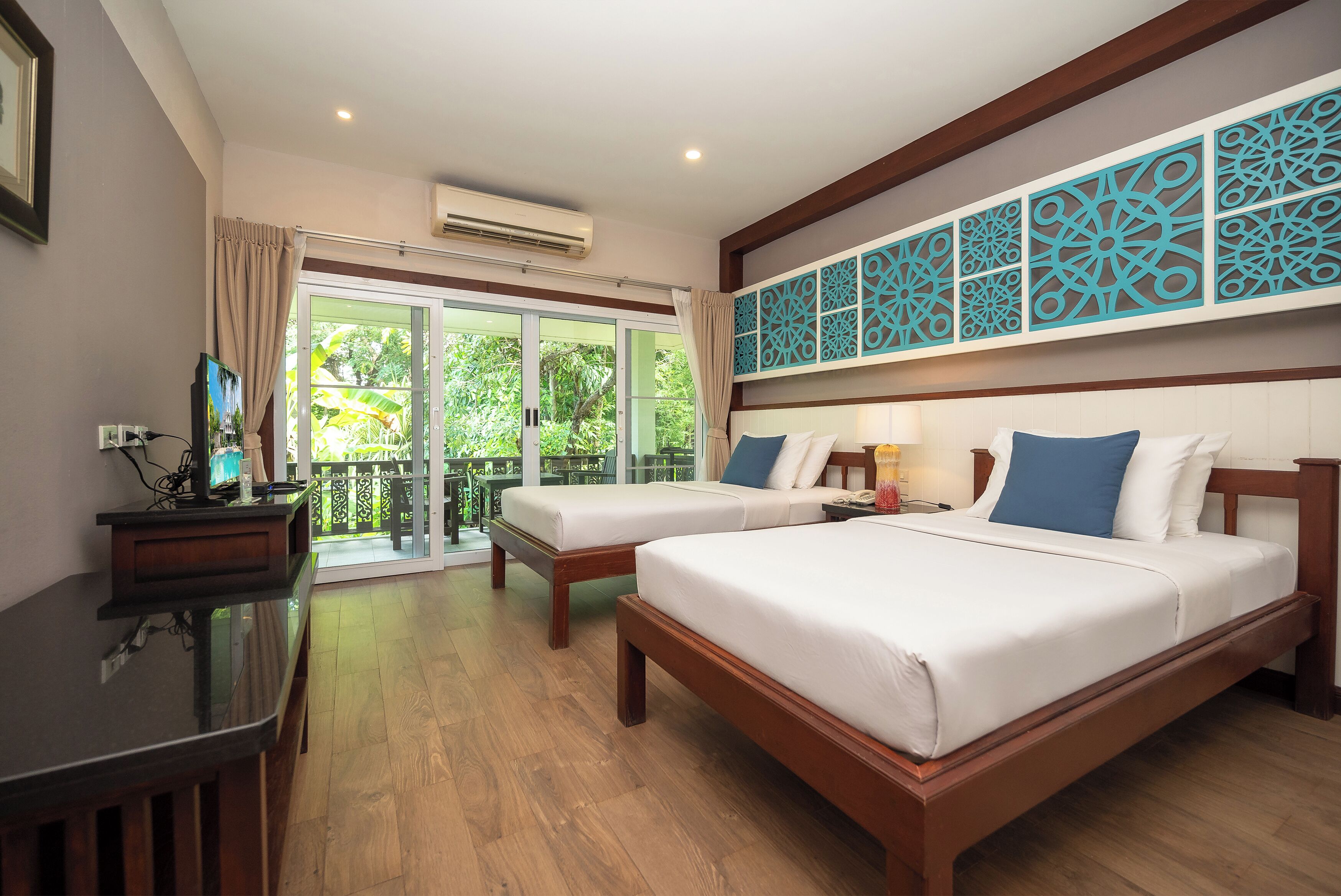 A hotel room with two beds, a desk, and a balcony with a view of greenery.