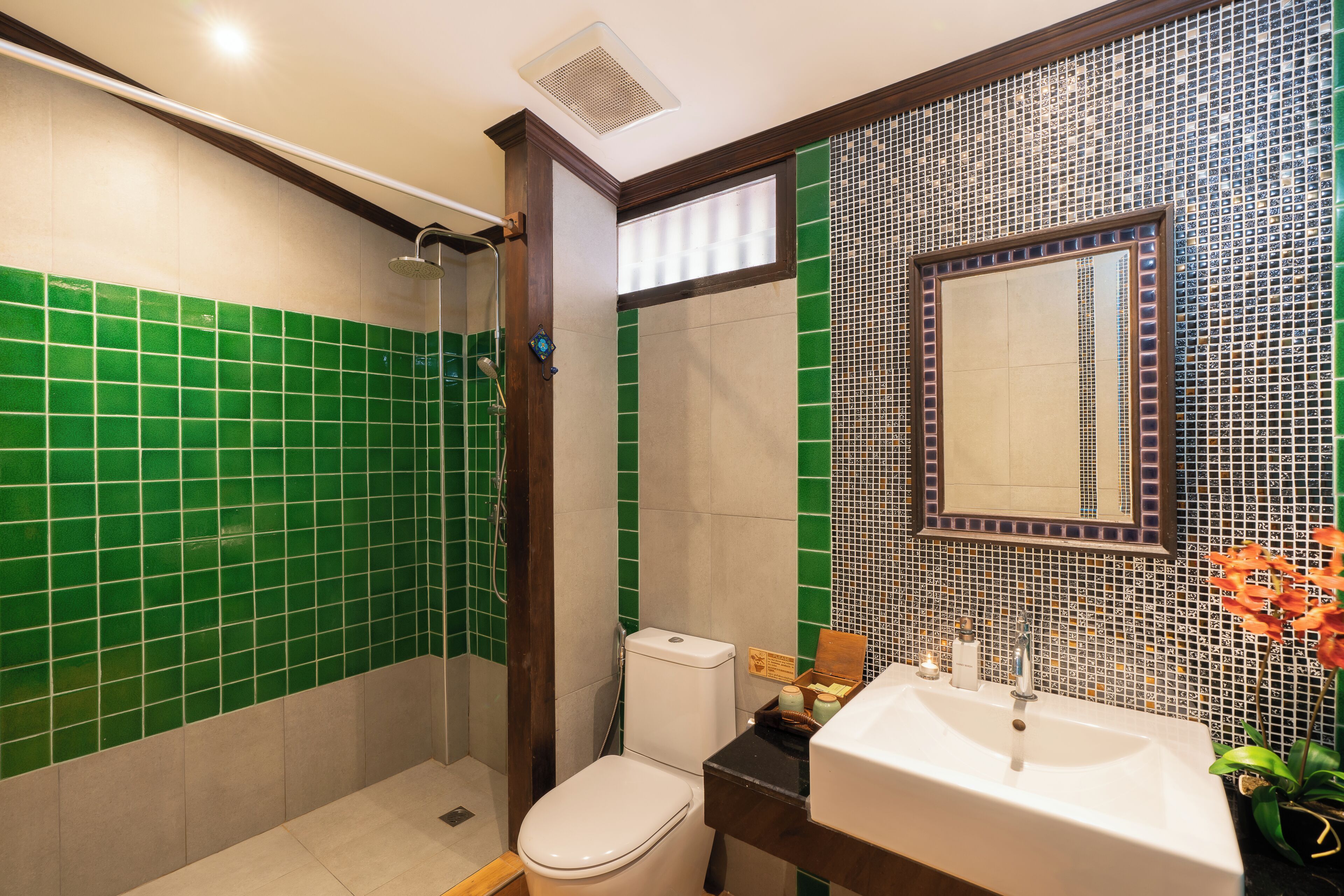 Een badkamer met groene en beige tegels, een wit toilet, een douche, een wastafel en een spiegel.