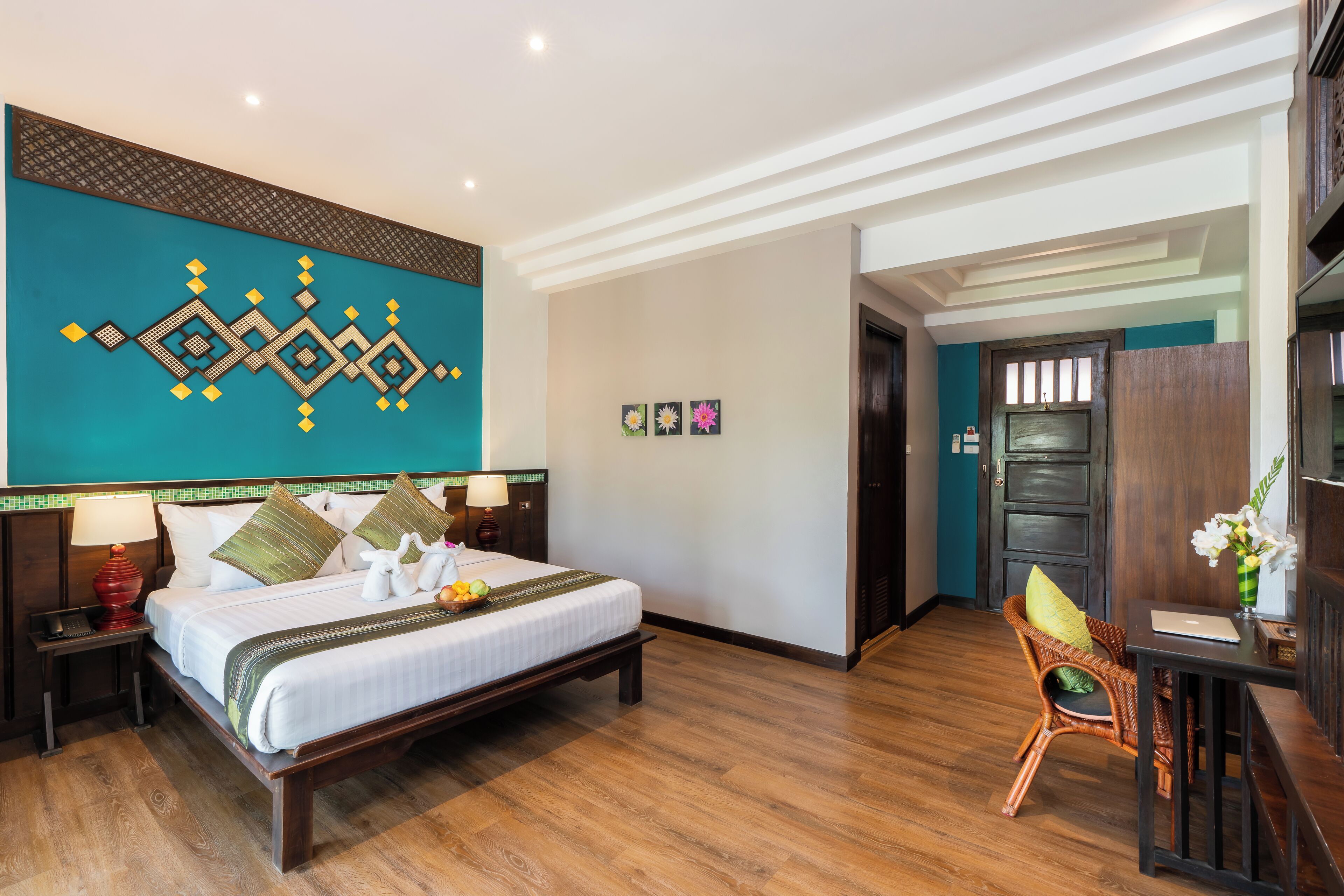 A hotel room with a large bed, a desk with a laptop, a wicker chair, and a decorative wall with geometric patterns.
