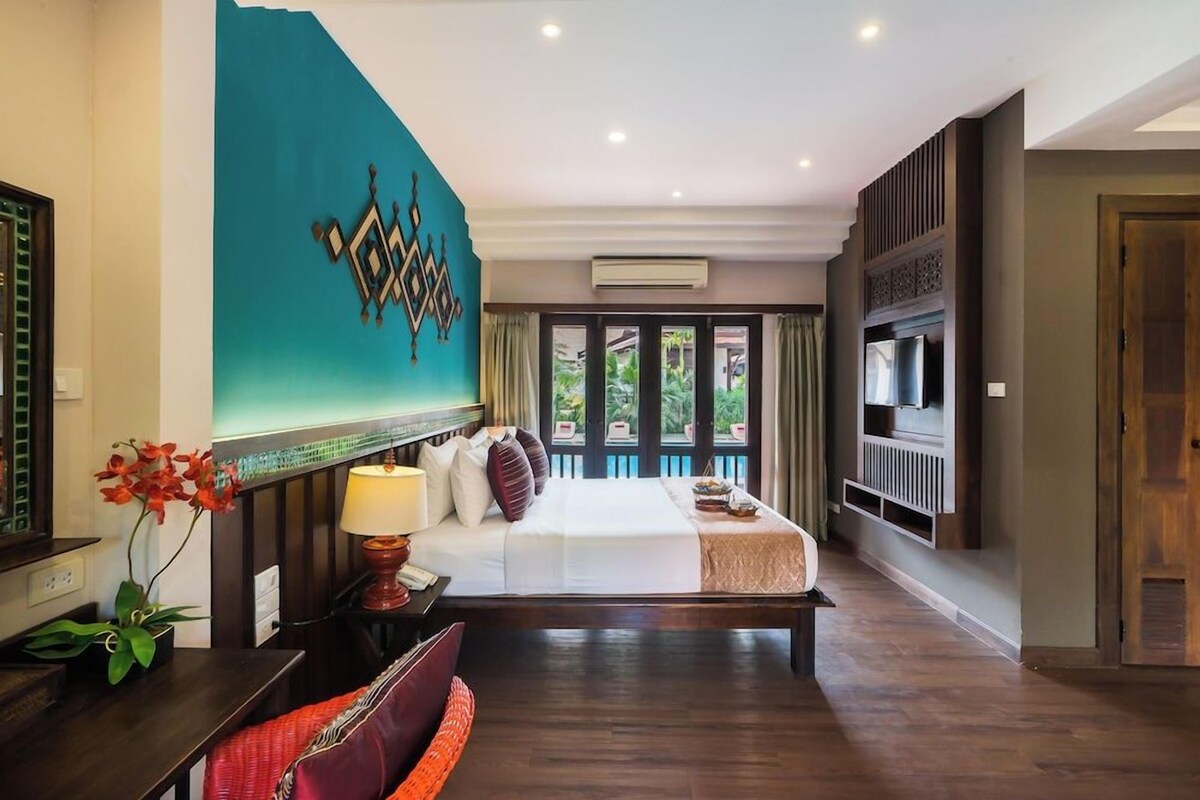 A hotel room with a bed, a TV, a desk, and a window with a view of greenery.