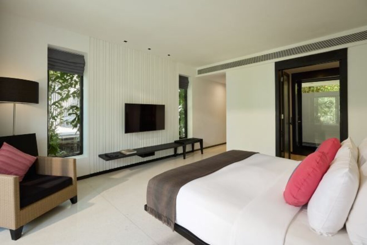 A modern hotel room with a large bed, a sofa, a TV, and a window with a view of greenery.