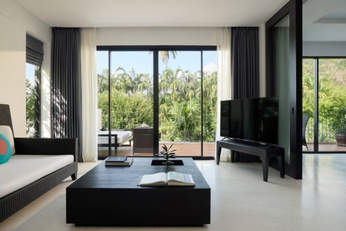 A modern living room with a large TV, a black coffee table, and a sofa. There is a balcony with a view of palm trees.