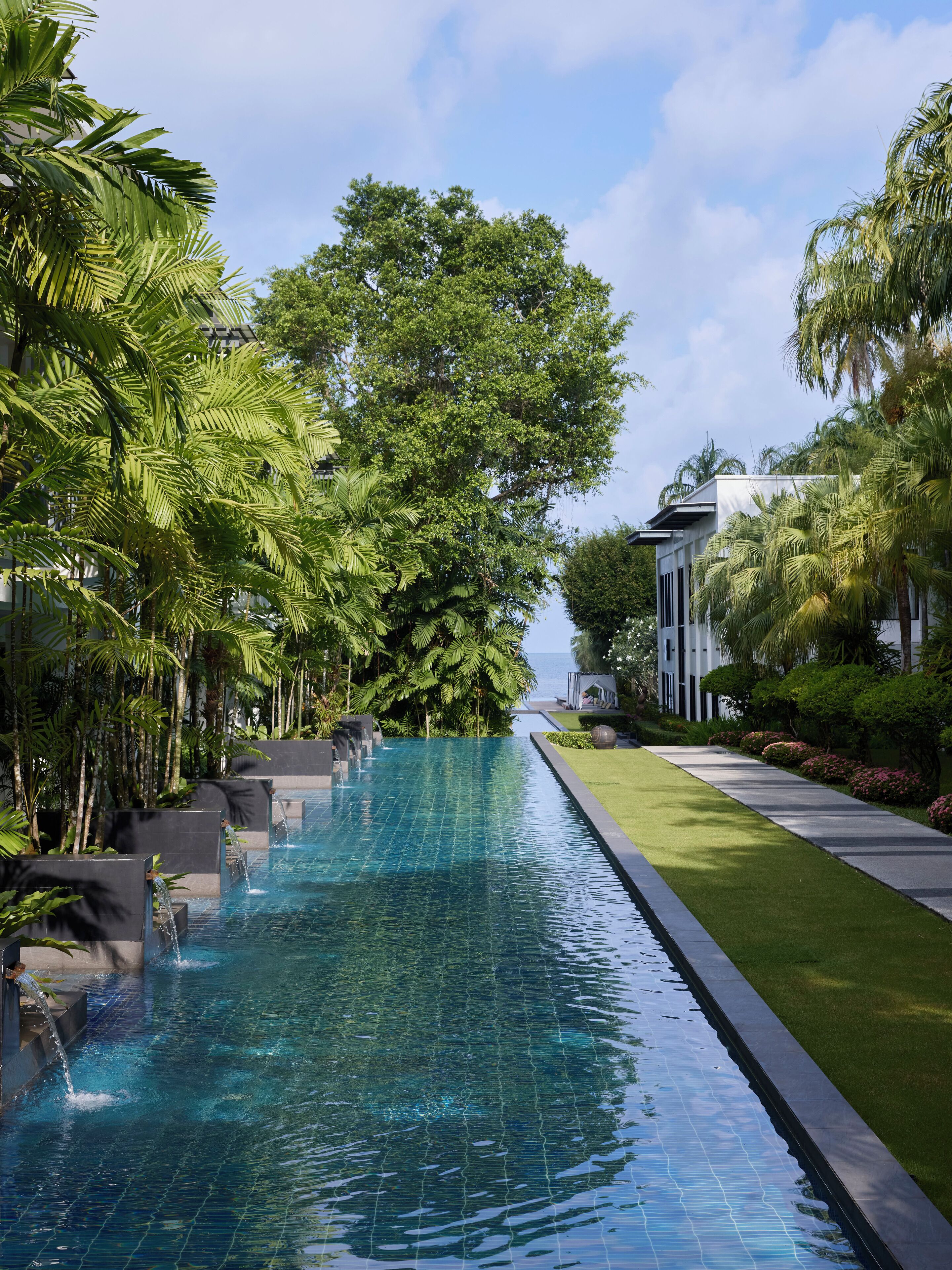 A long, narrow swimming pool with clear blue water, surrounded by lush greenery and palm trees.