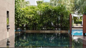 A poolside area with a view of a pool, surrounded by lush greenery and palm trees. There are two lounge chairs and a table with a book and glass.