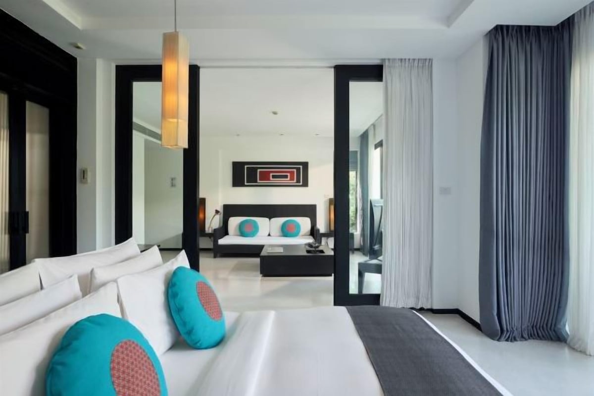 A modern hotel room with a large bed, a sofa, and a dining area. There is a view of the city through large windows.