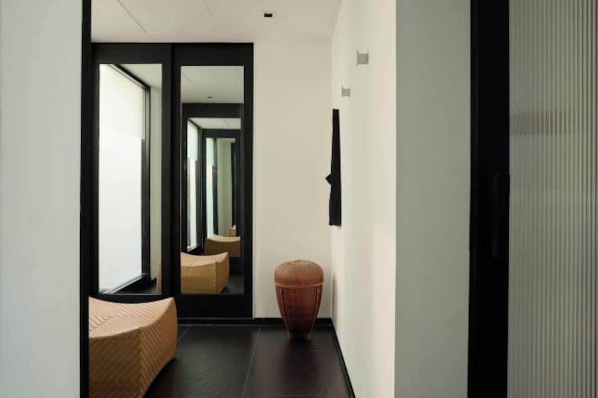 Un pasillo moderno de hotel con un espejo grande que refleja la habitación, un tapete con textura y un taburete tejido original.