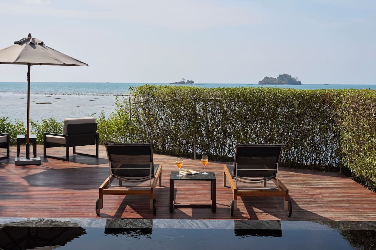 Terraza de madera con sillones y una mesa, con vista a un mar azul y unas islas lejanas.