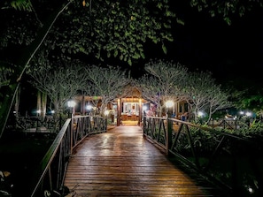 A wooden bridge leading to a building with a covered entrance, surrounded by trees and illuminated by lights.