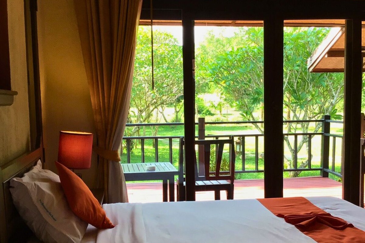 Habitación de hotel con dos camas, una ventana con cortinas, vista a zonas verdes y un balcón con mesa y sillas.