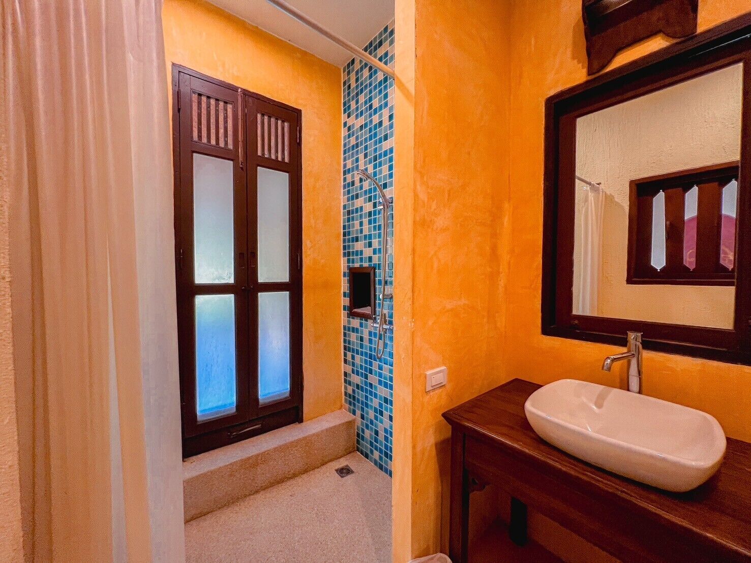 Un baño con puerta de madera, zona de ducha con azulejos y un lavabo blanco con espejo.