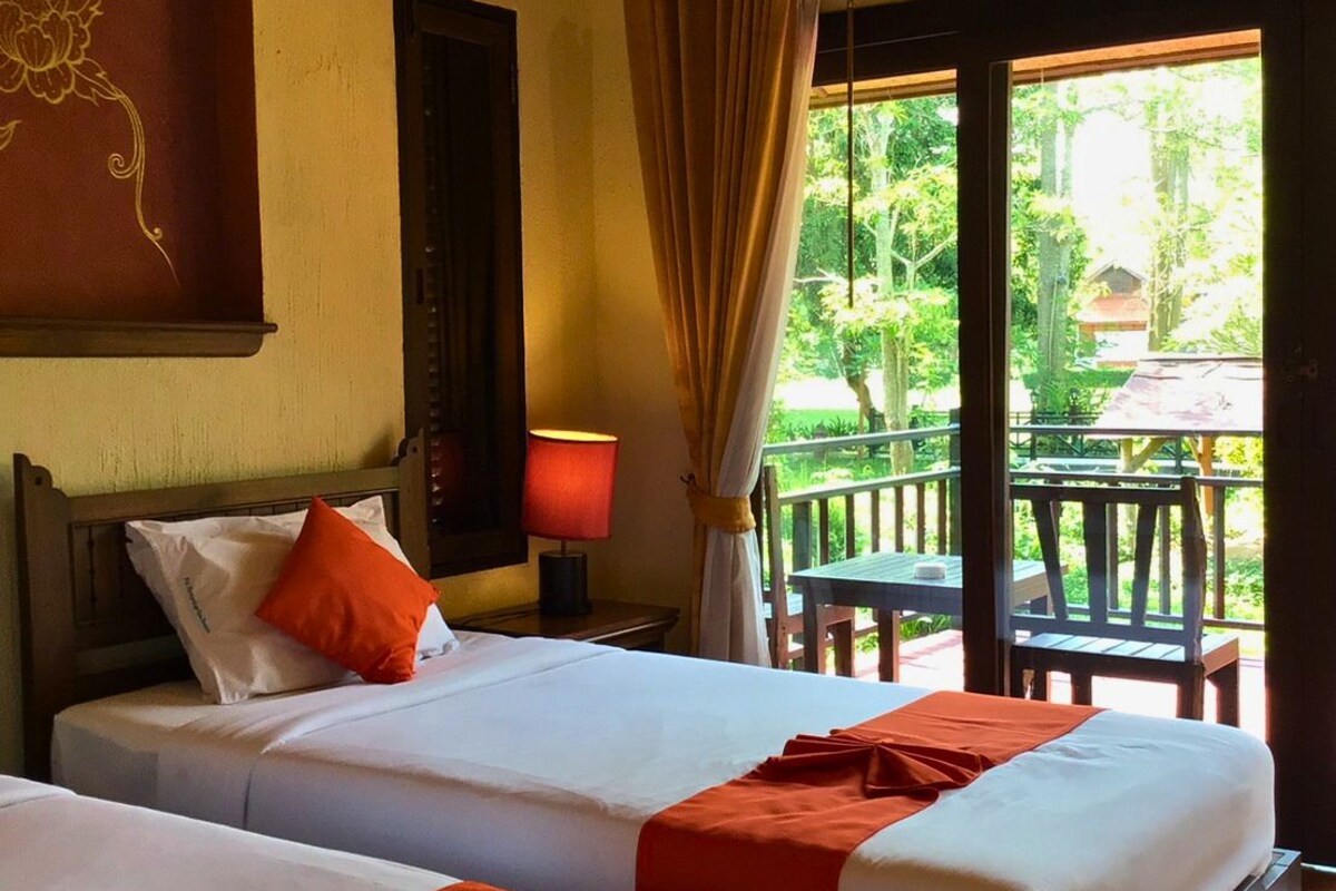 A hotel room with two beds, a nightstand, a lamp, and a view of a balcony with outdoor furniture.