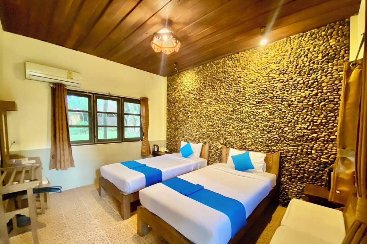 A room with two beds, a wooden ceiling, a stone wall, a window with curtains, and a small table with a chair.