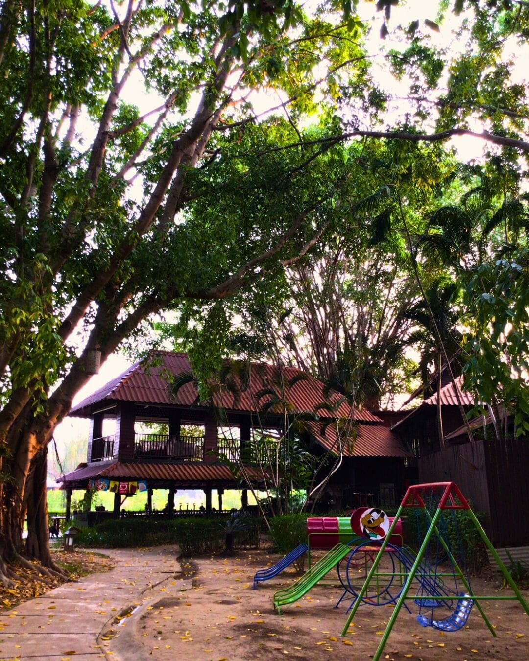 A playground with slides and swings, surrounded by trees and a building with a red roof.