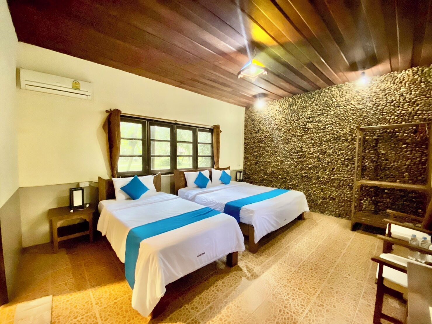 Una habitación con dos camas, una pared de piedra, techo de madera y una pequeña mesa con botellas.