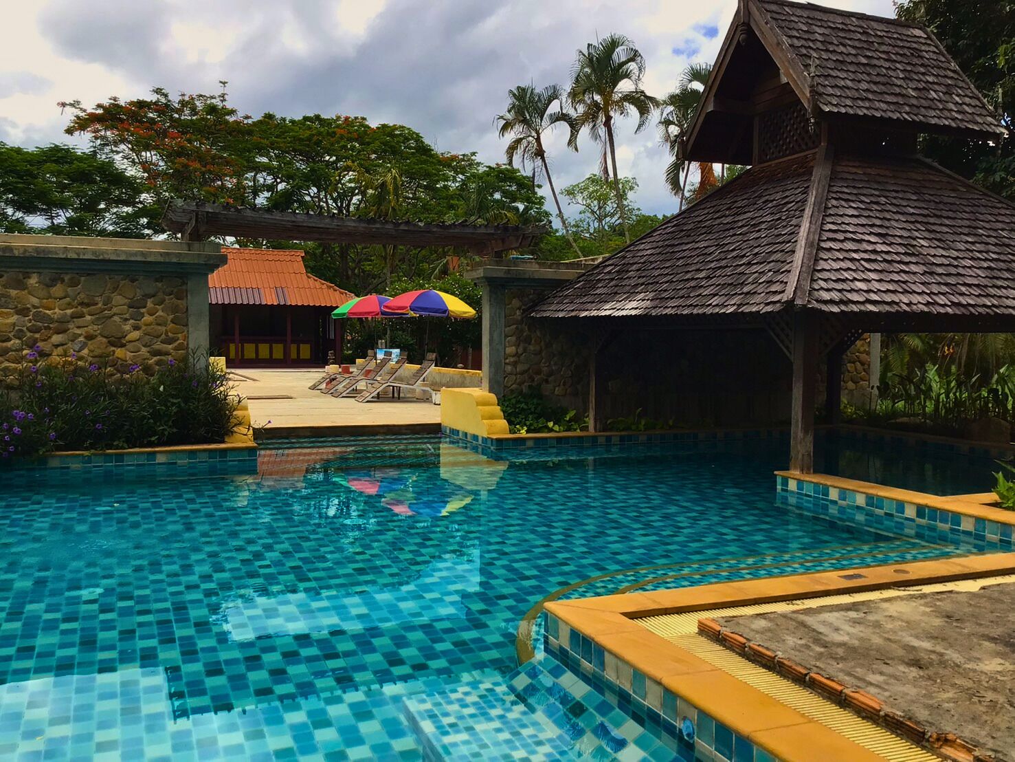 Una piscina con bordes de azulejos, un área de descanso con tumbonas y una estructura con techo de paja.