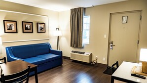 A room with a blue sofa, a wooden table with a chair, a lamp, and a window with curtains. - Whitman Motor Lodge (Huntington Station)