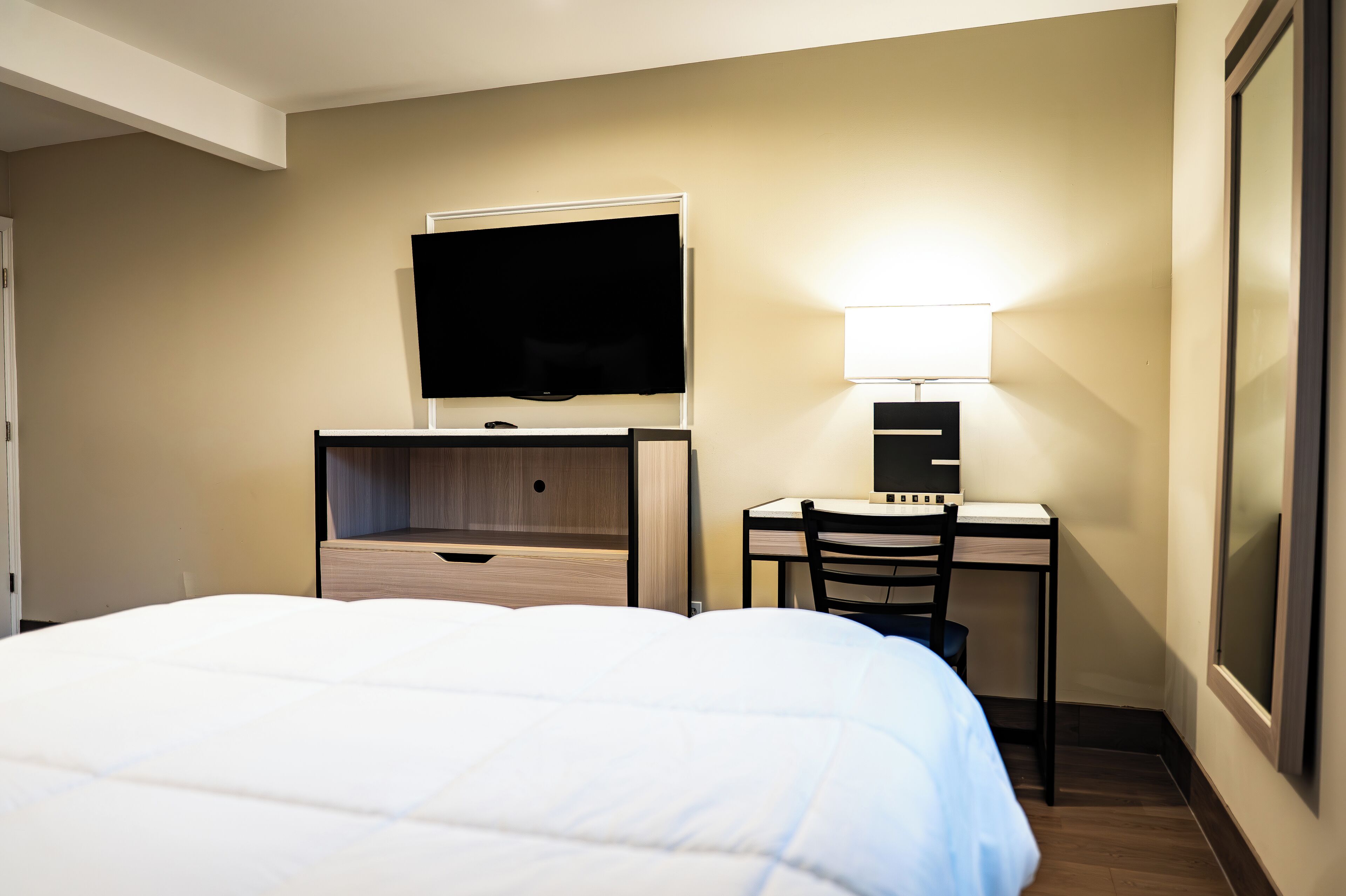 Quarto de hotel com uma cama, uma escrivaninha com cadeira, uma televisão e uma luminária.