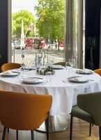 Ein runder Esstisch für vier Personen, gedeckt mit weißen Tischdecken, Besteck und Weingläsern. Die Stühle sind orange und grün gepolstert.