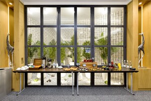 A buffet table with various food items and a coffee station, set against a backdrop of patterned curtains and a view of greenery.