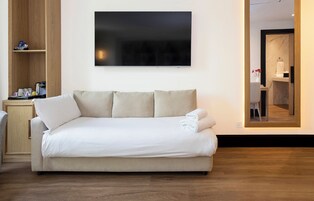 Un sofá de tonos neutros con una manta blanca y almohadas, un mueble de madera con artículos de tocador, y un espejo grande enmarco que refleja un baño.