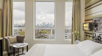 A hotel room with a bed, a chair, a small table, a nightstand, a lamp, and a view of the city skyline through large windows.