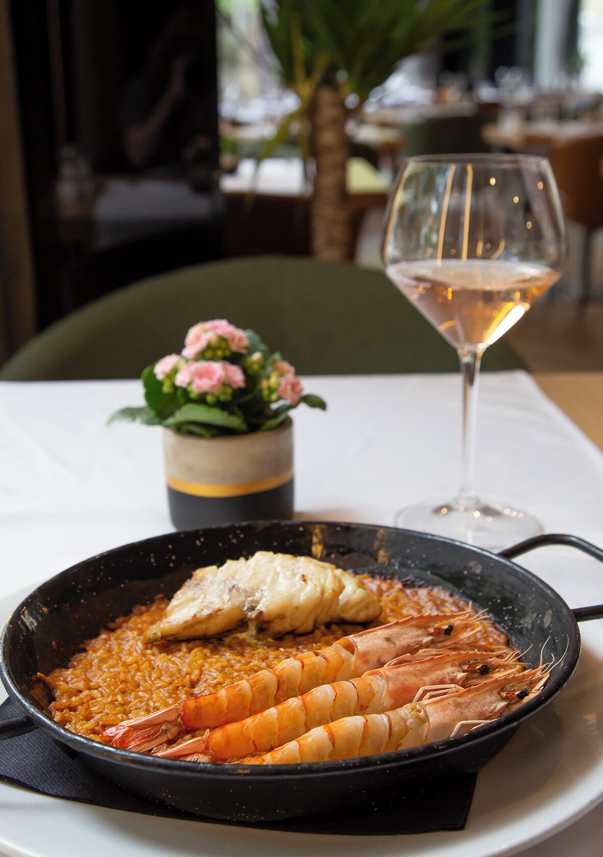 Plato de mariscos con camarones y pescado, una copa de vino blanco y una pequeña planta en maceta sobre una mesa.