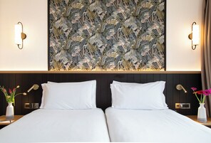 Camera d'hotel con un grande murale di foglie tropicali, due letti bianchi, comodini con lampade e un tavolo da notte con un vaso di fiori.