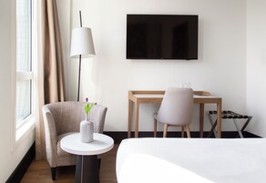 Ein Hotelzimmer mit einem Bett, einem Schreibtisch, einem Sessel, einem Fernseher, einer Lampe und einer Vase mit Blumen.