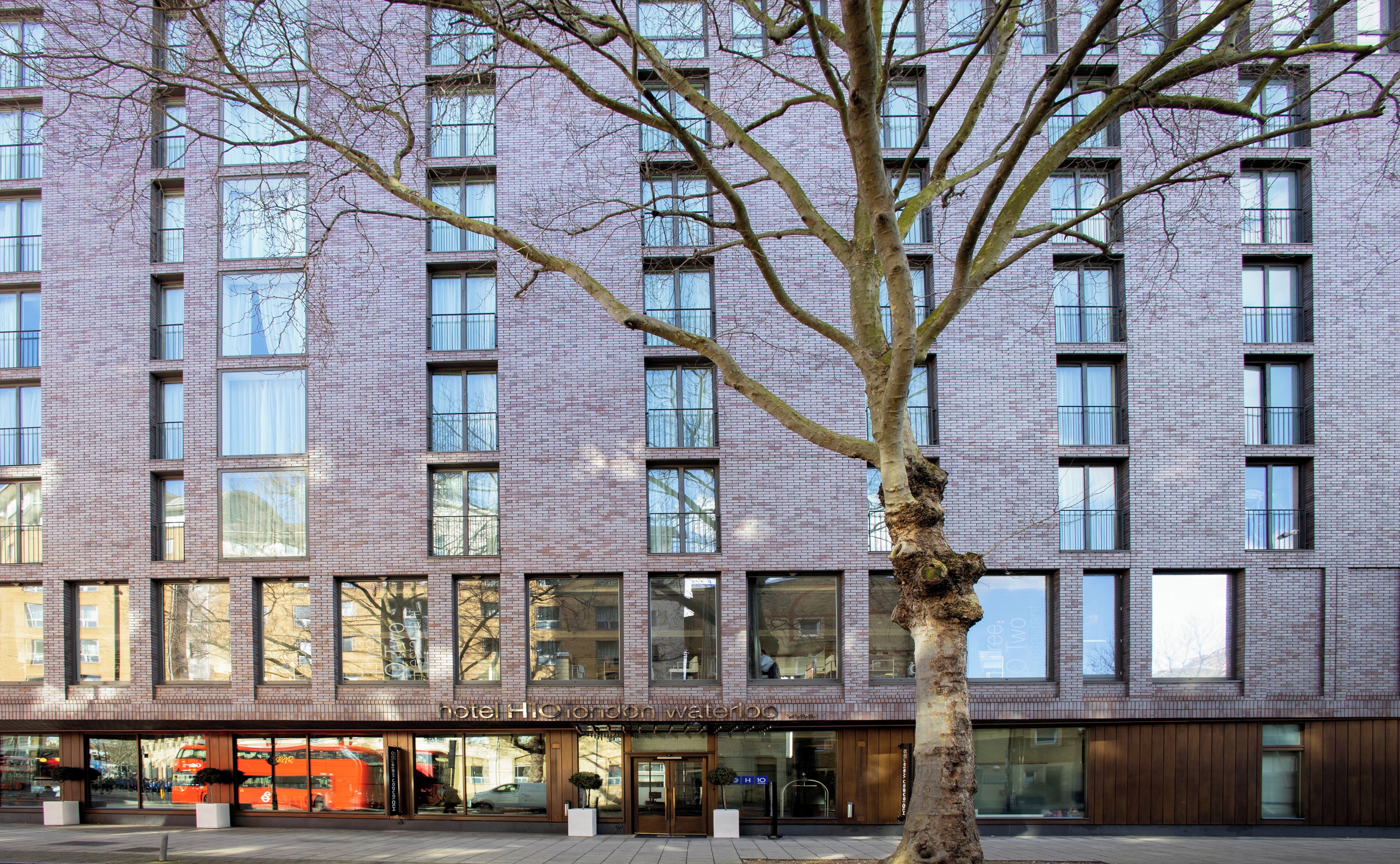 A modern hotel building with a brick facade, large windows, and a tree in front.