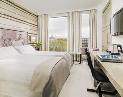 A hotel room with two beds, a desk with a chair, a TV, and a view of the cityscape.