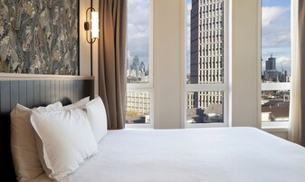 A hotel room with a large bed, a city view, a wall-mounted light fixture, and a patterned wallpaper.