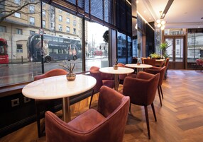 Un café moderno con mesas redondas y sillones cómodos, grandes ventanales que ofrecen vistas a la calle y un autobús rojo que pasa.