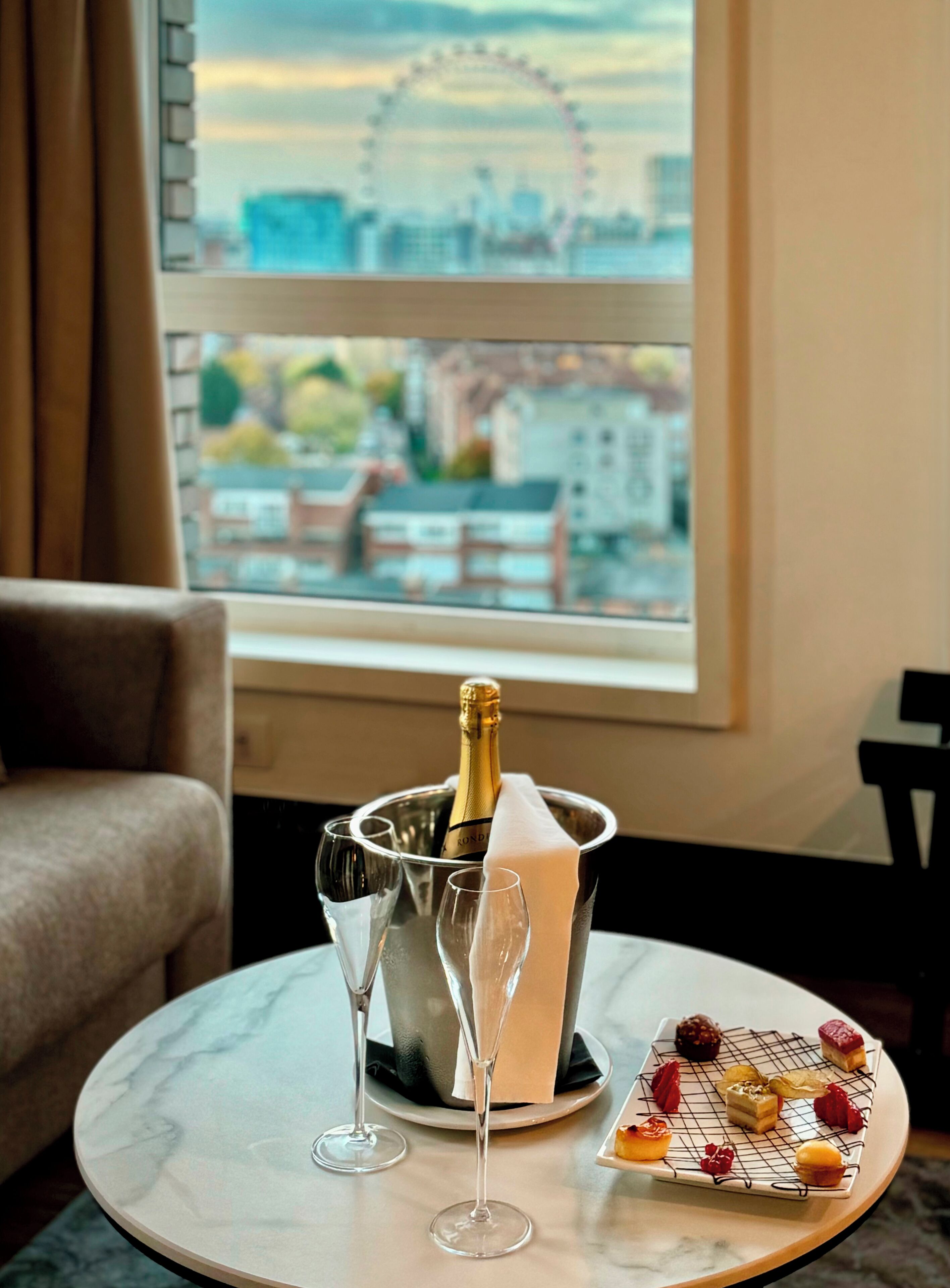 A room with a view of a cityscape, a table with a champagne bottle, two glasses, and a dessert tray.