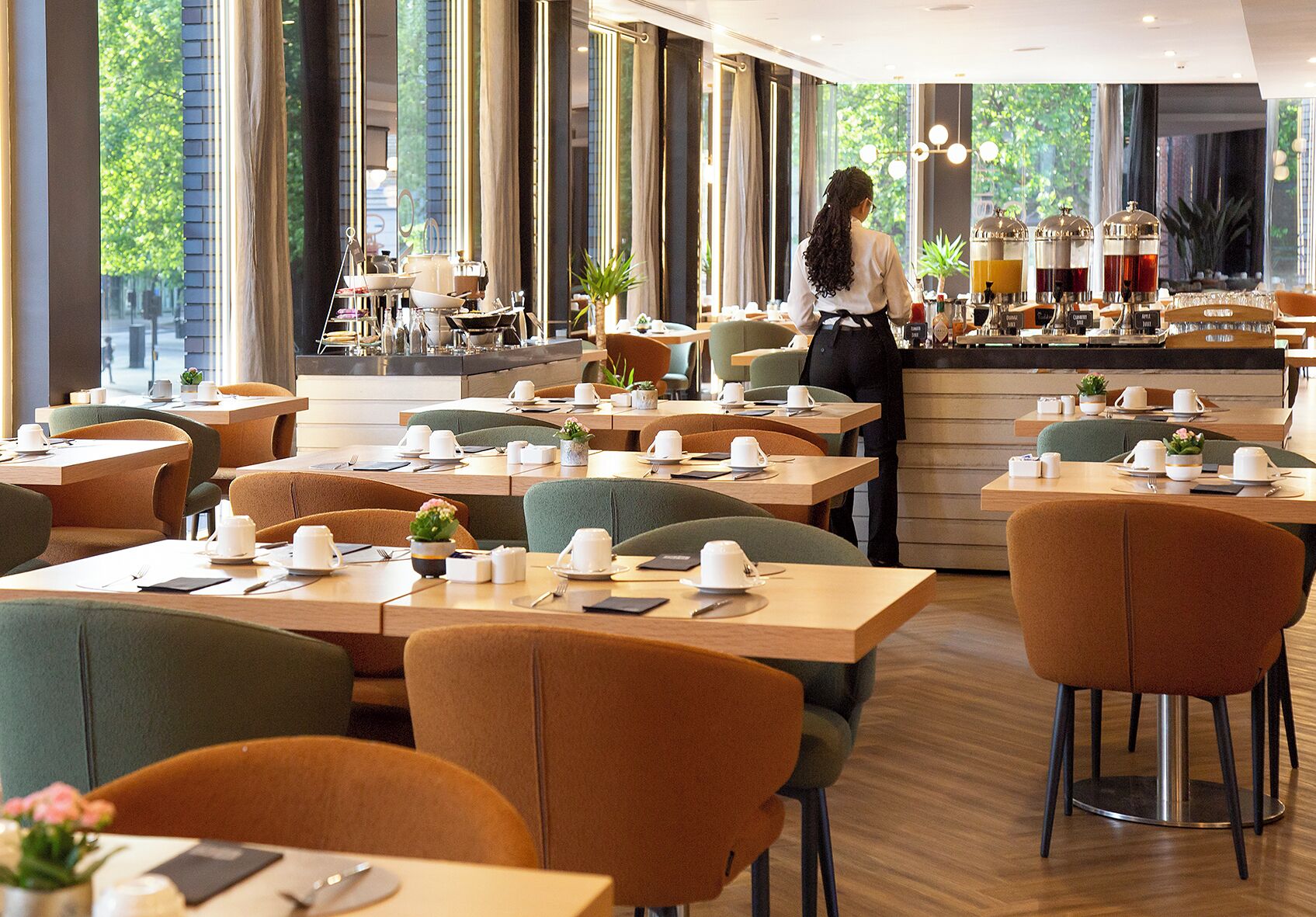 Un café moderne avec des tables préparées pour le petit-déjeuner, un bar où l’on sert des boissons et de grandes fenêtres donnant sur l’extérieur.