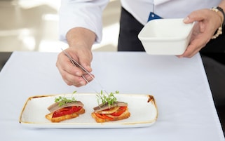 A person serving food from a white bowl onto a rectangular plate with two pieces of toast topped with vegetables and fish.