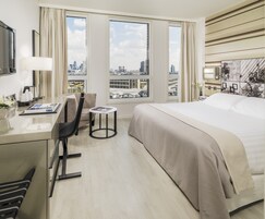 Habitación de hotel con cama, escritorio, silla, una mesita y vistas de la ciudad a través de amplios ventanales.