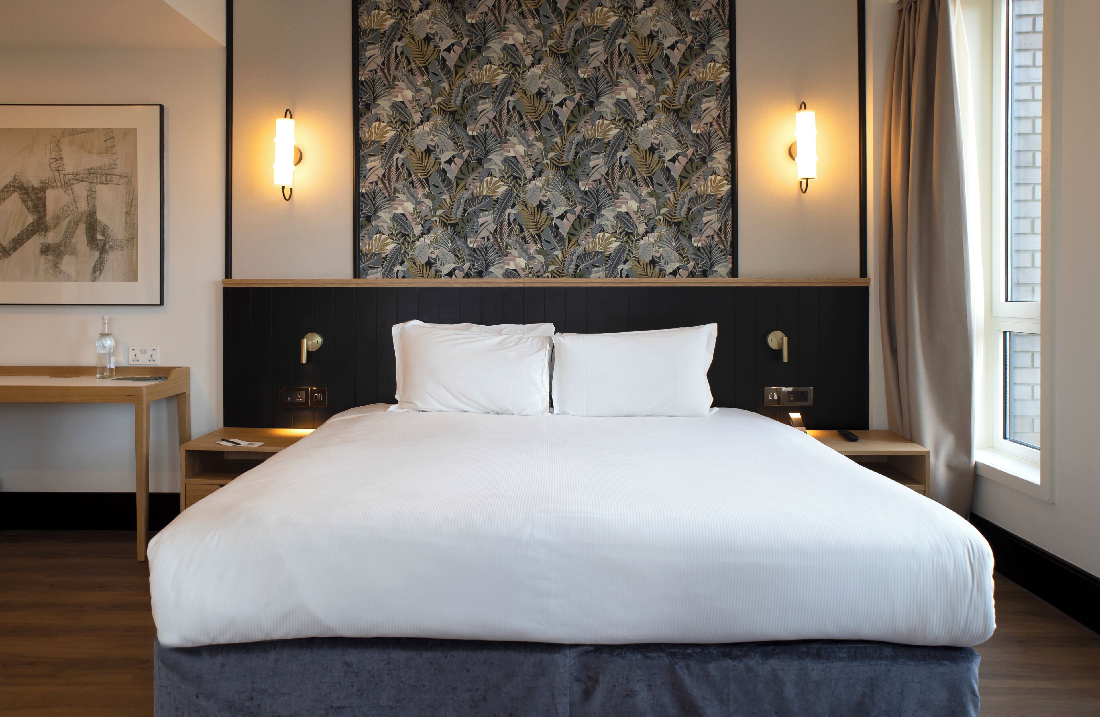 A hotel room with a large bed, two bedside tables, a wall-mounted artwork, and a window with curtains.