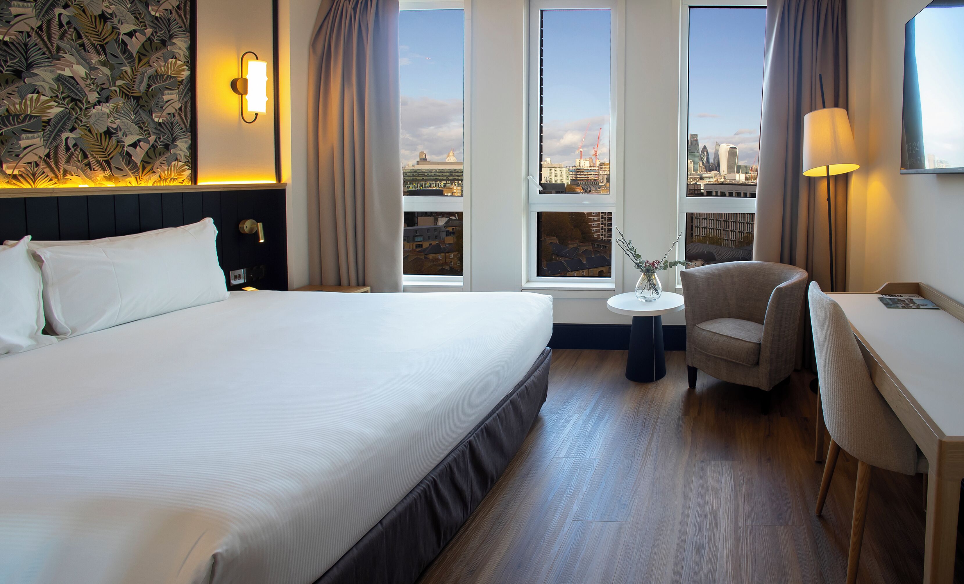 Camera d'albergo con un letto grande, una sedia, un tavolino e vista sulla città attraverso ampie finestre.