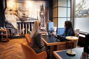 Zwei Personen sitzen an einem Holztisch mit einem Computer und einer Lampe in einem Zimmer mit einem großen Fenster und Wandgemälde eines berühmten Gebäudes.