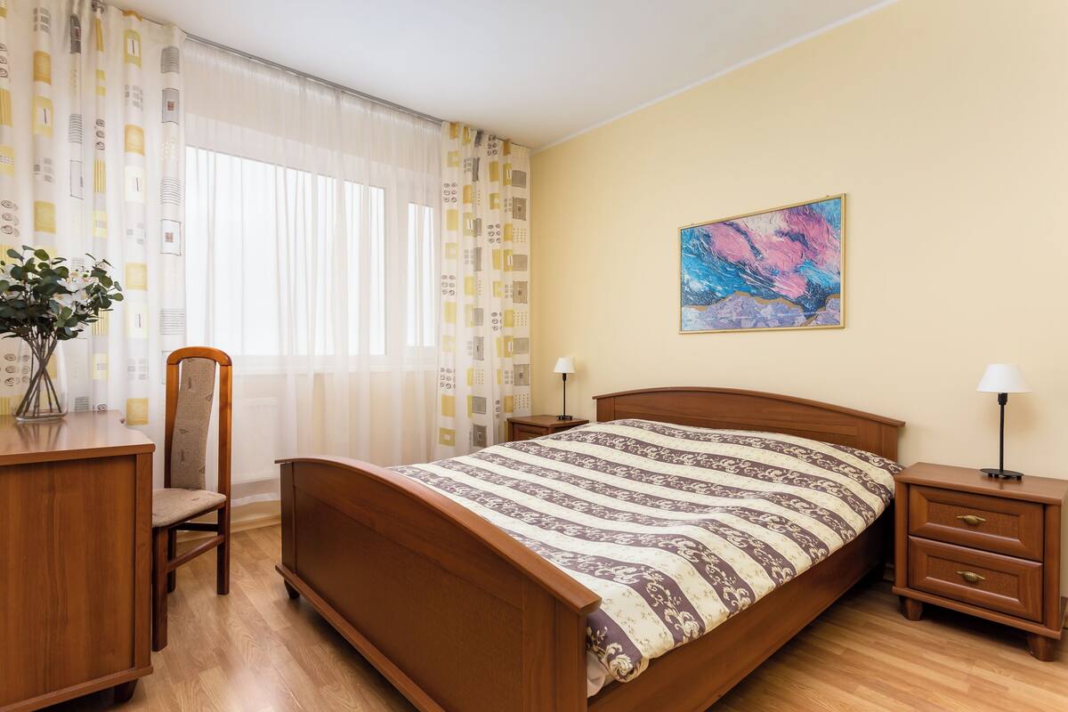 A bedroom with a wooden bed, a desk, a chair, a nightstand, and a lamp.