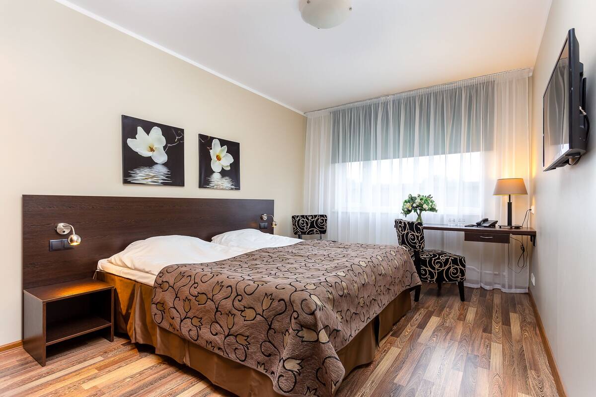 A hotel room with a wooden floor, a bed with a patterned bedspread, a desk with a chair, a TV, and artwork on the wall.