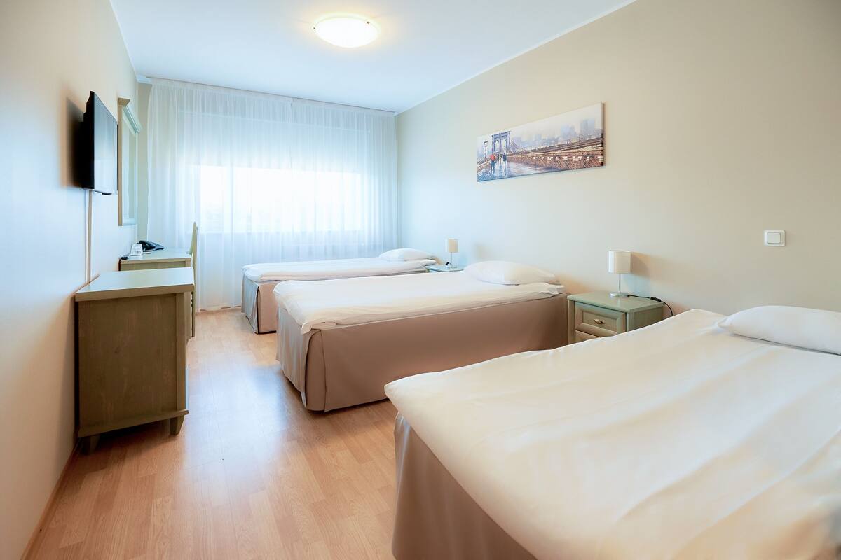 A hotel room with three beds, a desk, a chair, a TV, and a painting on the wall.