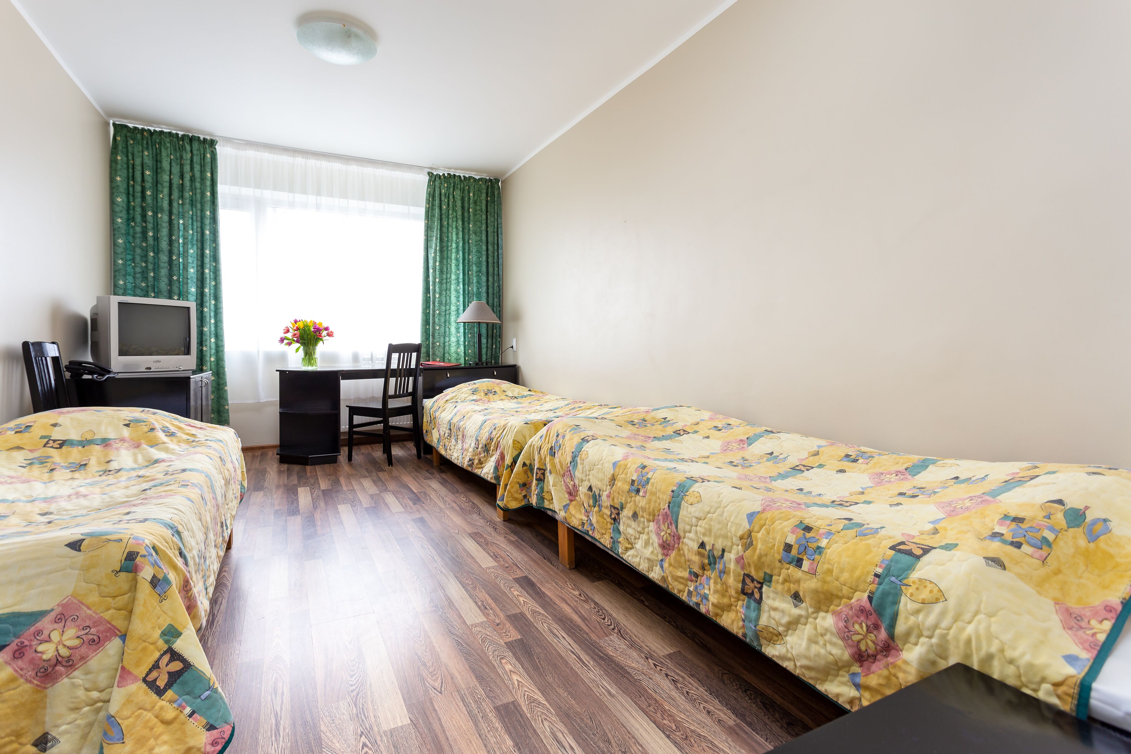 Ein Zimmer mit zwei Betten, einem Schreibtisch, einem Stuhl, einem Fernseher und einem Fenster mit Vorhängen.