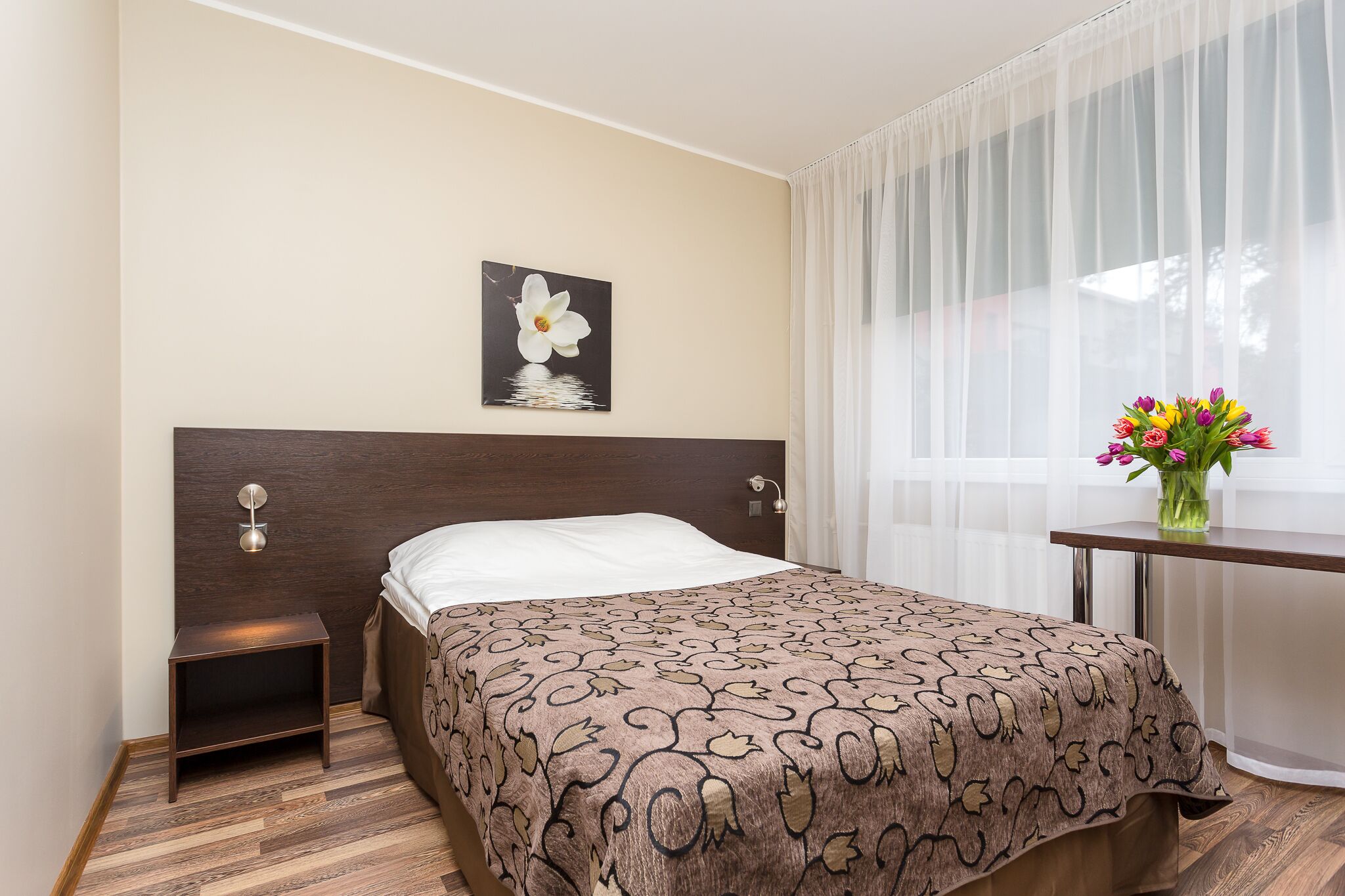 A hotel room with a bed, a nightstand, a wall-mounted lamp, a window with sheer curtains, and a vase with flowers.