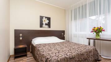 A hotel room with a bed, a nightstand, a wall-mounted lamp, a window with sheer curtains, and a vase with flowers.