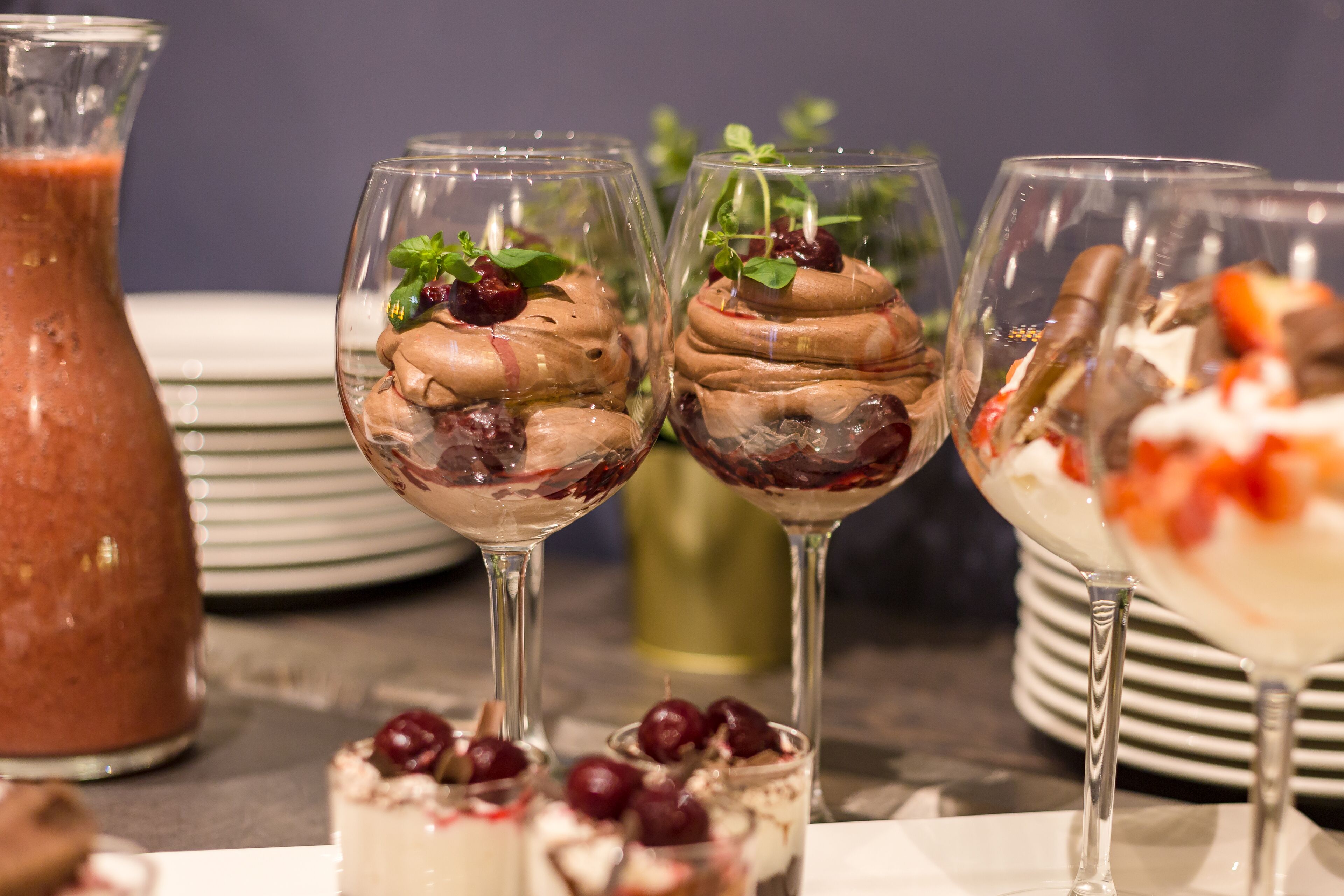 Tre dessertglas fyllda med lager på lager av olika desserter och körsbär, en kanna med en röd dryck och en hög med vita tallrikar.